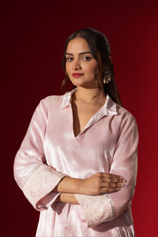 Closeup view of a woman stands in a reflective pink velvet co-ord set with a long tunic and draped pants, against a vibrant red backdrop.