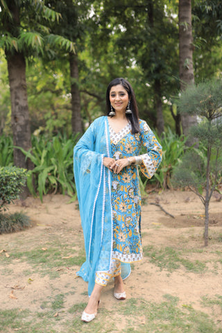 A woman stands outdoors in a park-like setting wearing a traditional handblock printed outfit. The outfit consists of a light blue kurta with intricate yellow and white floral patterns, paired with a solid light blue dupatta and matching light blue trousers.