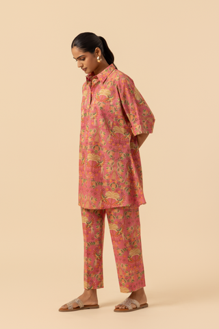 Side view of a woman stands in a vibrant pink and floral-patterned co-ord set, featuring a loose shirt and matching pants, set against a neutral background.