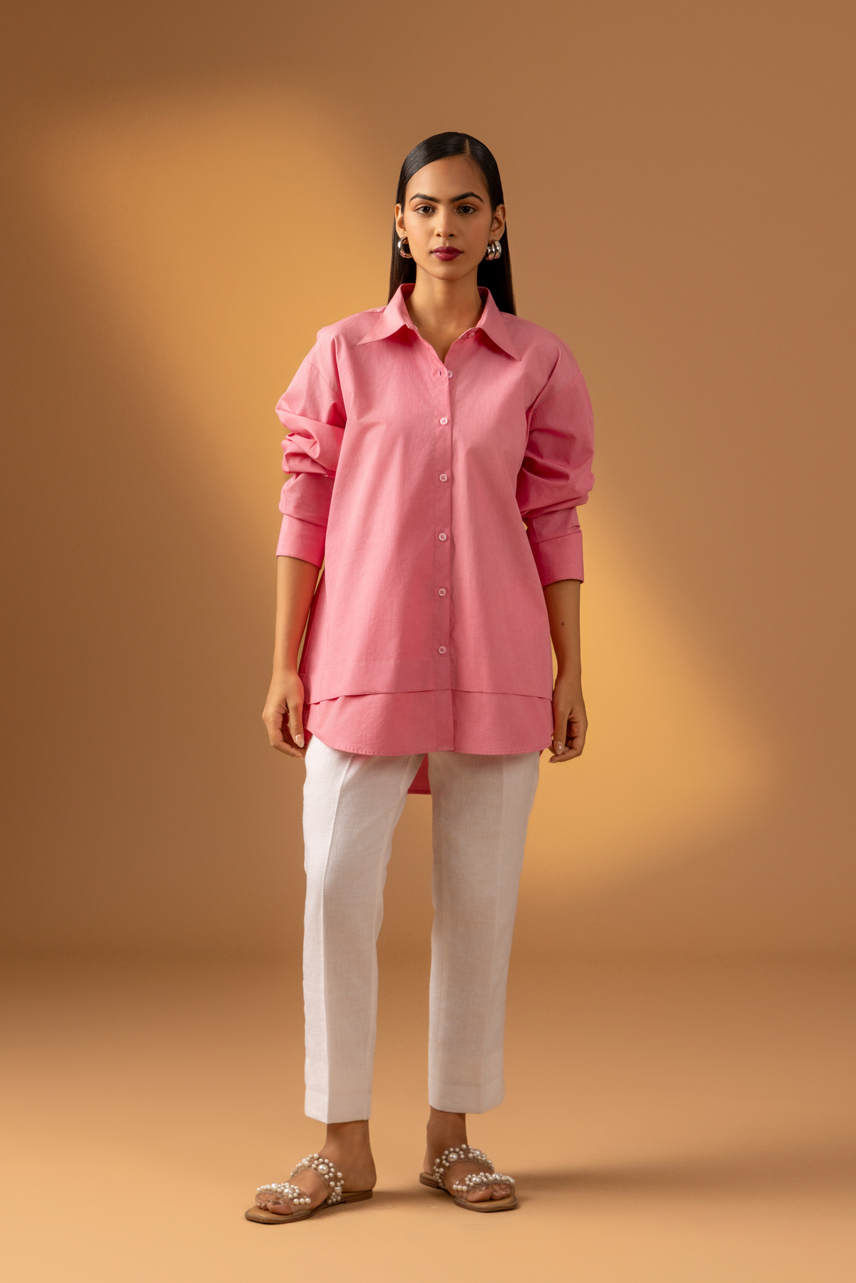 A Woman wearing a pink shirt paired with white trousers, and sandals against a warm, neutral background.