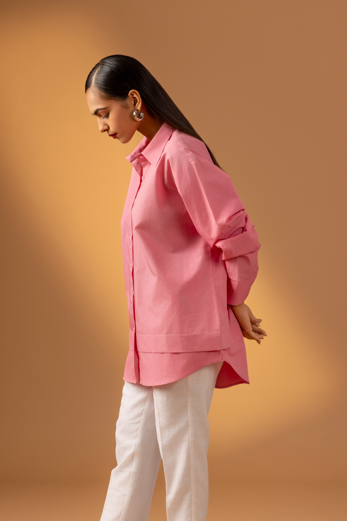 Side view of a woman wearing a pink shirt paired with white trousers, and sandals against a warm, neutral background.