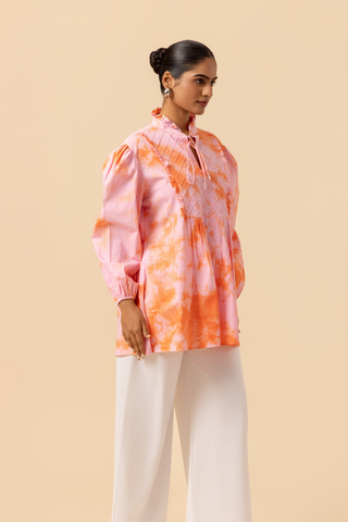 Right side view of a model wears a vibrant light pink & orange tie-dye shirt dress with puffed sleeves, paired with white wide-leg trousers and stylish sandals.