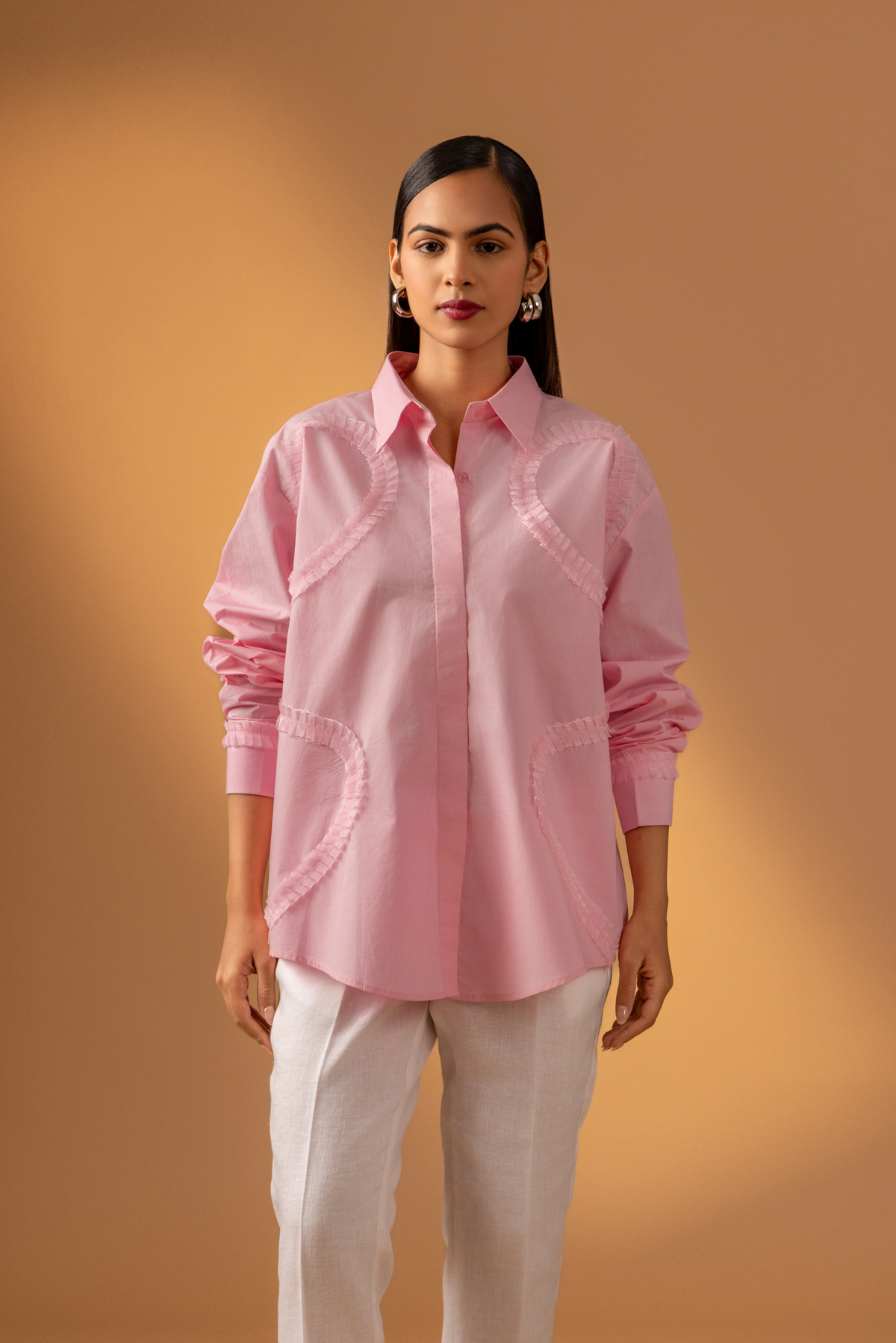 Closeup view of a woman stands against a warm, neutral backdrop, wearing a pink oversized shirt, white trousers, and stylish sandals.