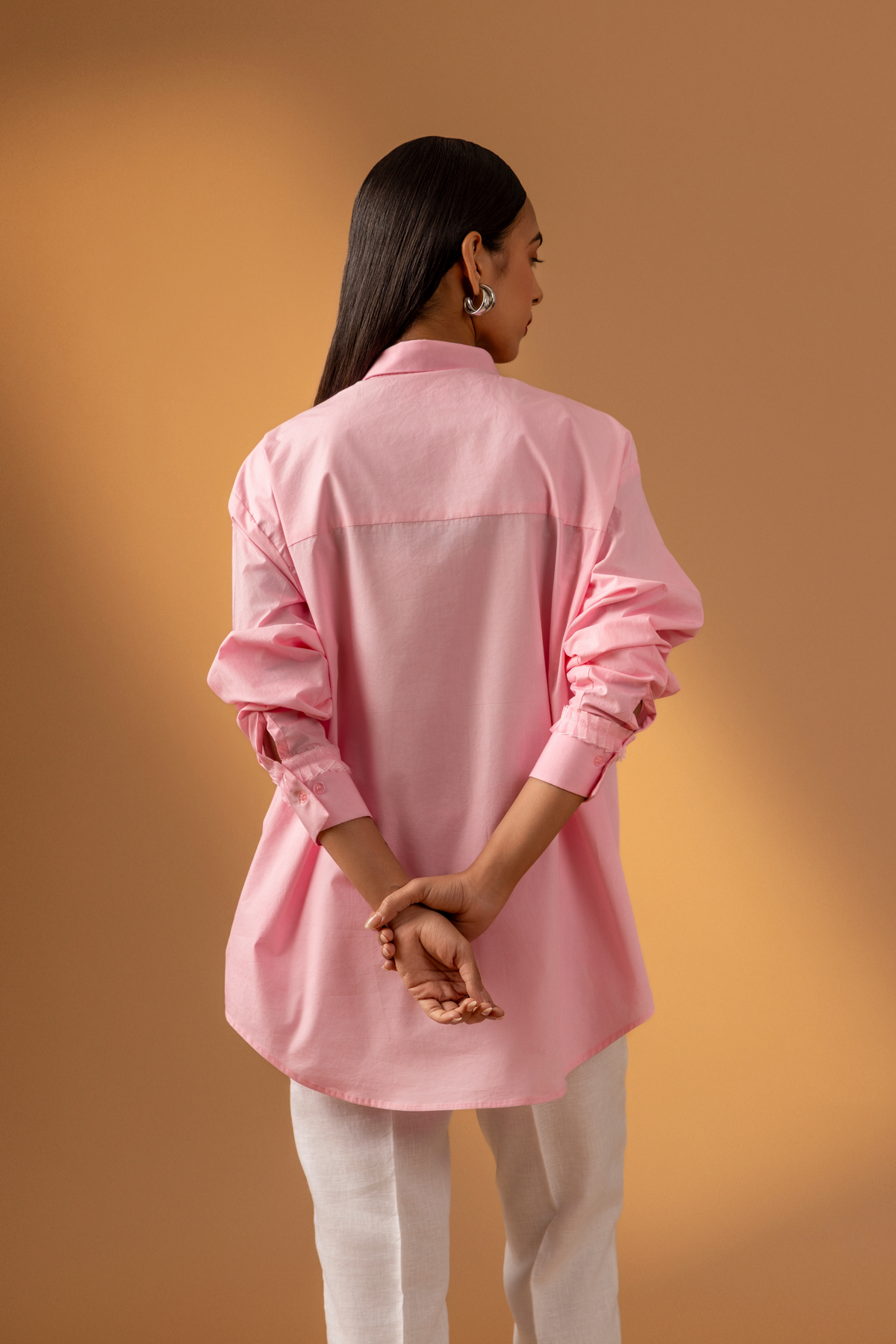 Back view of a woman stands against a warm, neutral backdrop, wearing a pink oversized shirt, white trousers, and stylish sandals.