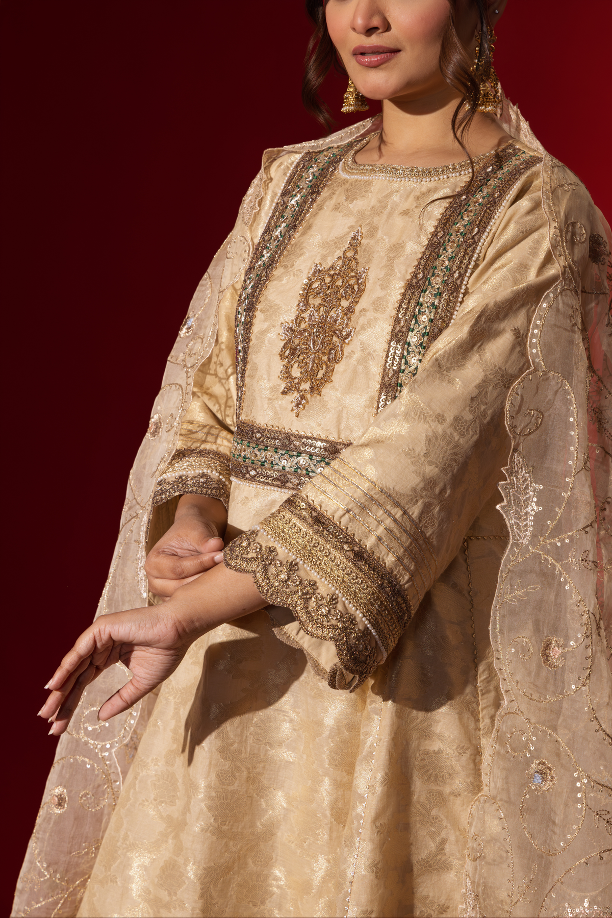 Closeup view of a woman's hand wearing an elegant cream embroidered sequin brocade kurta set with gold accents and a sheer dupatta, set against a rich red background