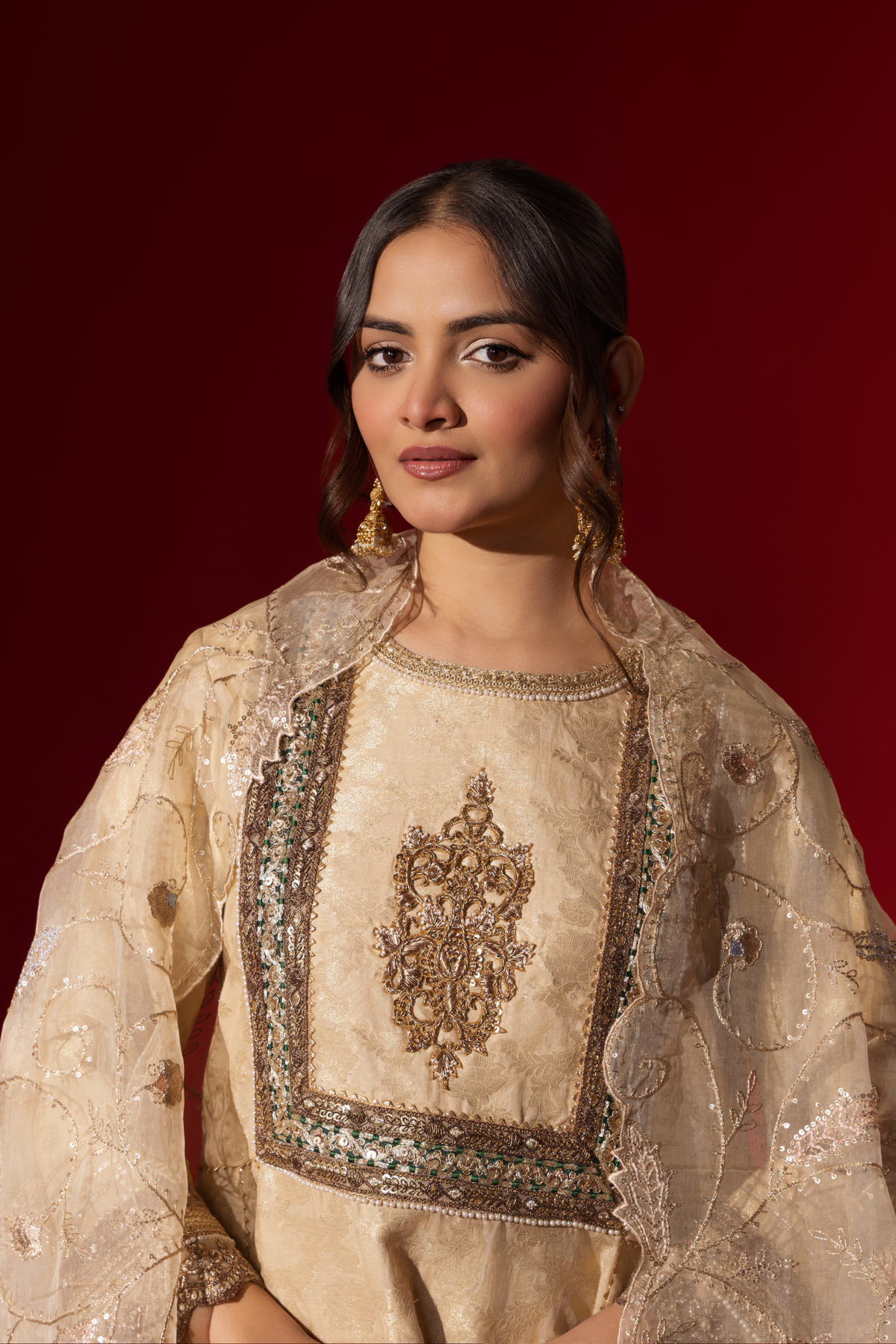 Closeup view of a woman wearing an elegant cream embroidered sequin brocade kurta set with gold accents and a sheer dupatta, set against a rich red background