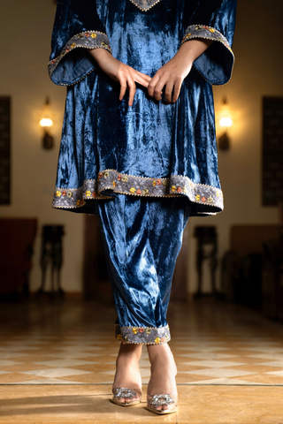 A woman stands elegantly in a royal blue velvet traditional outfit with embellished cuffs, showcasing her stylish shoes in a softly lit space.