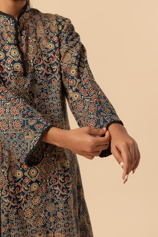 Closeup view of a model's hand wearing a long, patterned ajrakh print co-ord set which has tunic and loose pants, showcasing intricate designs in dark blues and golds against a neutral background.