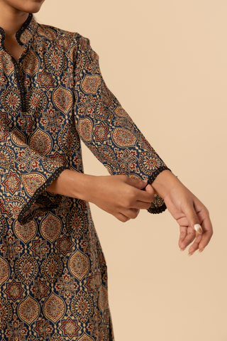Closeup view of a model's hand  stands in a patterned ajrakh print co-ord set which has brown kurta and matching loose pants against a soft beige background, showcasing traditional ethnic wear.