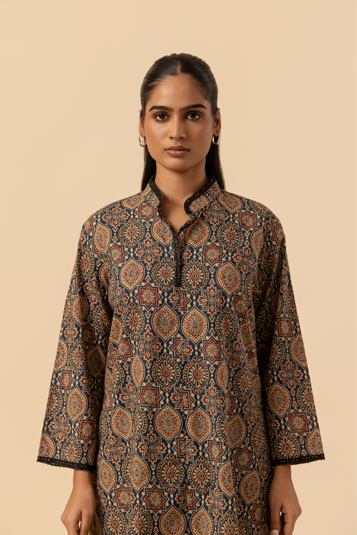 Closeup view of a model stands in a patterned ajrakh print co-ord set which has brown kurta and matching loose pants against a soft beige background, showcasing traditional ethnic wear.