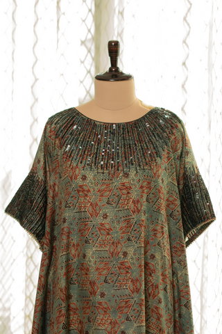 Closeup view of green and rust printed kaftan with intricate pleated detailing and sequin embellishments on the neckline, displayed on a mannequin against a softly lit curtain backdrop.