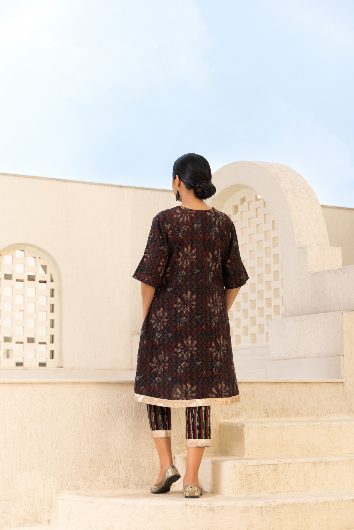 Back view of a Model dressed in a brown printed kurta set with flower intricate patterns, paired with striped trousers, styled in a serene outdoor setting