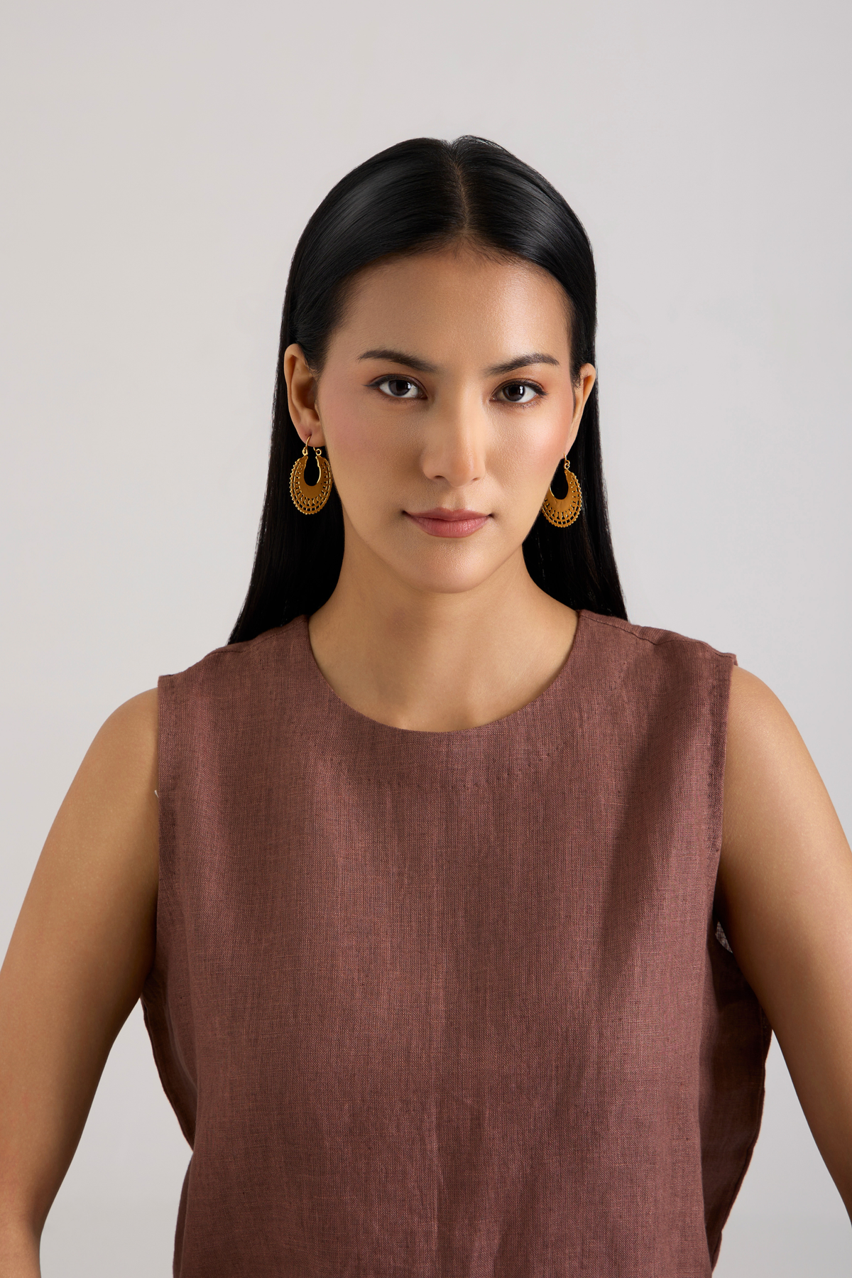 A woman with long black hair wearing a sleeveless dusty rose-brown top and gold hoop earrings, photographed against a plain light gray studio background.