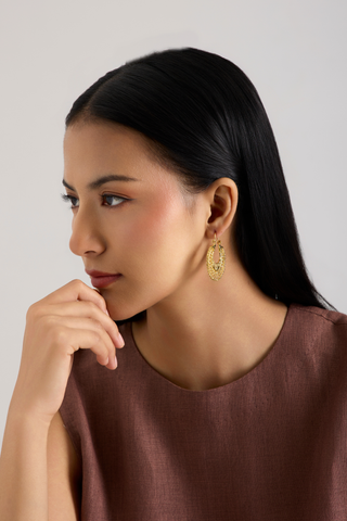 A woman with long black hair wearing a brown top and a gold-toned dangle earring, shown in a close-up portrait against a light gray background.