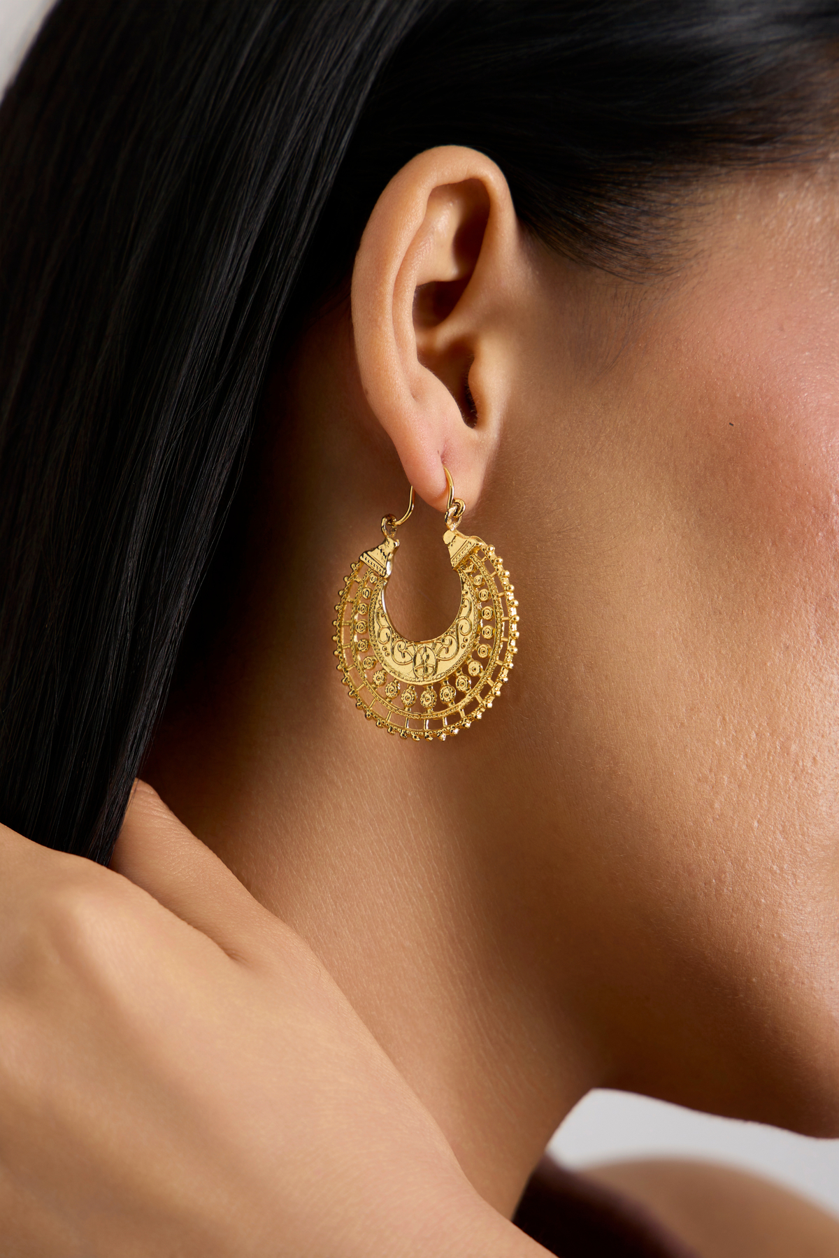 A close-up side profile of a model wearing a large, ornate gold crescent hoop earring with intricate filigree detailing.