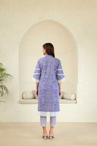 Back View of a woman wearing a purple traditional outfit with intricate white embroidery and lace detailing, paired with matching pants, standing in front of an arched alcove in a neutral-toned interior.