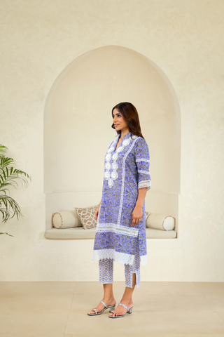 A woman is dressed in a purple traditional outfit with intricate white embroidery and lace detailing, paired with matching pants, standing in front of an arched alcove in a neutral-toned interior.