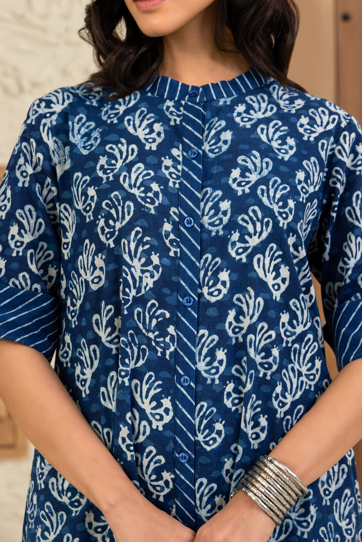 Closeup view of a model dressed in a blue and white printed co-ord set outfit with striped cuffs, standing against a wooden and textured beige background.
