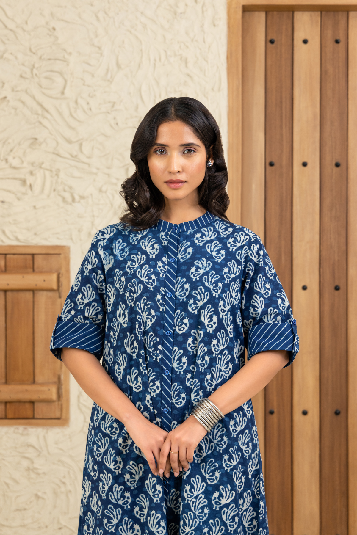 Closeup view of a woman wearing a blue and white printed co-ord set outfit with striped cuffs, standing against a wooden and textured beige background.