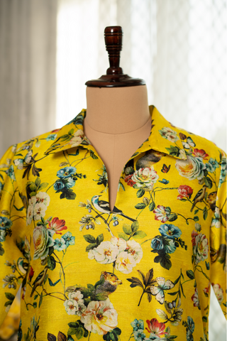 Closeup View of a  vibrant yellow floral dress displayed on a mannequin stand, set in a room with soft natural lighting, white curtains, and a decorative side table with yellow flowers.