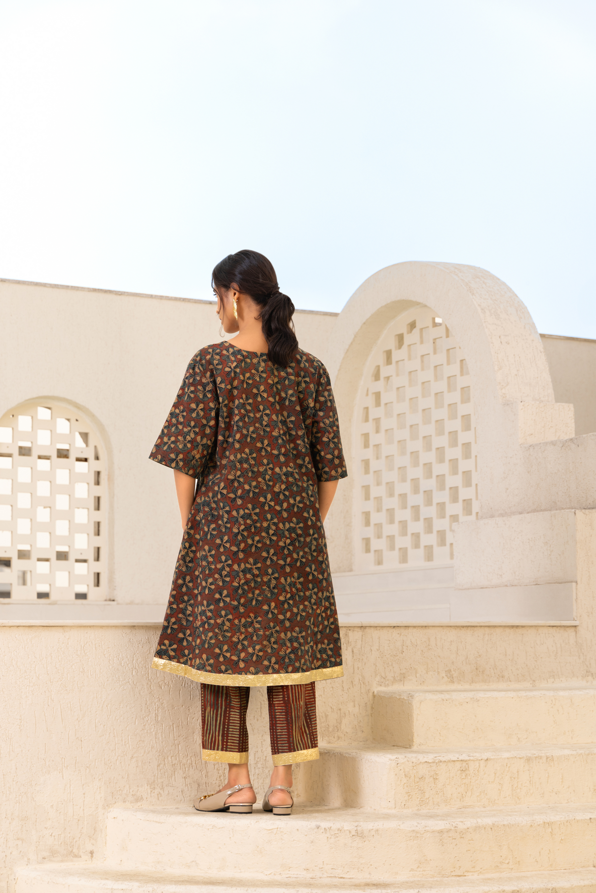 Back View of a model showcasing a brown floral printed kurta set with gold accents, paired with striped trousers, styled against an elegant outdoor backdrop.