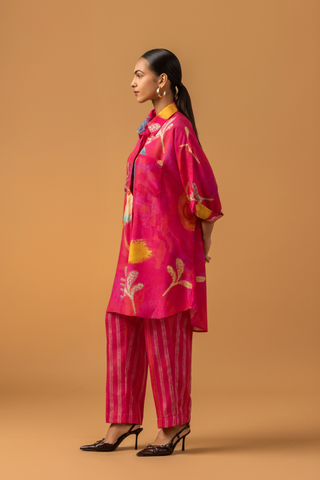 A model in a vibrant pink tunic with floral patterns co-ord set, paired with striped pink pants and stylish heels, against a warm, neutral background.