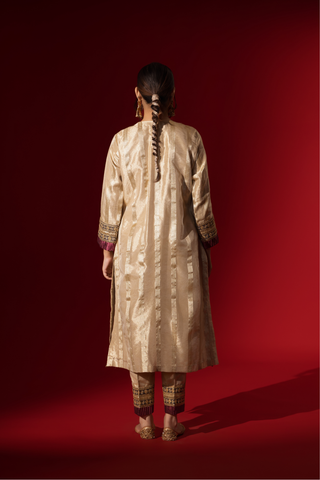 Back view of a Woman in a shimmering gold brocade kurta set stands confidently against a rich red backdrop, showcasing intricate patterns and styling.