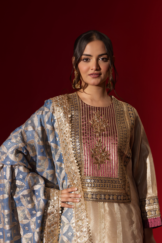 Closeup view of a Woman in a shimmering gold brocade kurta set stands confidently against a rich red backdrop, showcasing intricate patterns and styling.