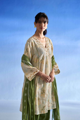 Closeup view of a woman modeling a Indian linen kurta set consisting of a light beige floral kurta, a green dupatta, and green trousers, set against a blue gradient studio background.