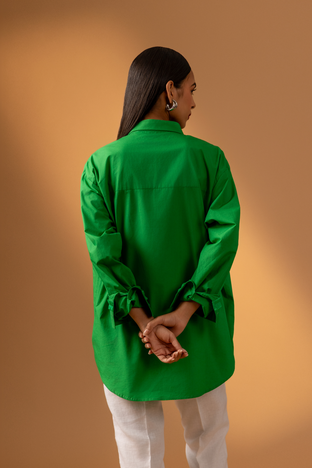Back view of a model wears a vibrant green shirt with unique detailing, paired with white pants, against a soft brown background.