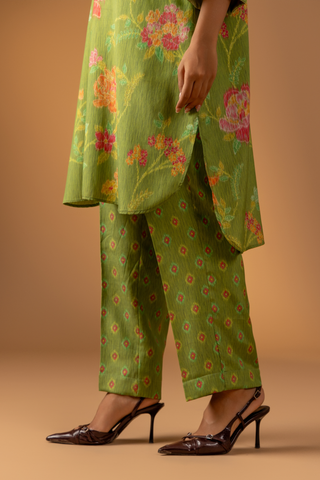 A close-up of a woman wearing a vibrant green outfit with floral patterns and chic brown heels against a soft, warm background.