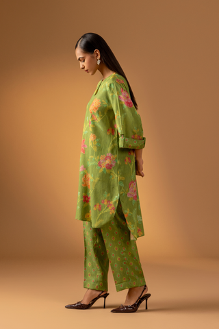 Side view of a model stands in a vibrant green co-ord set with floral patterns, paired with matching trousers, against a warm beige background.