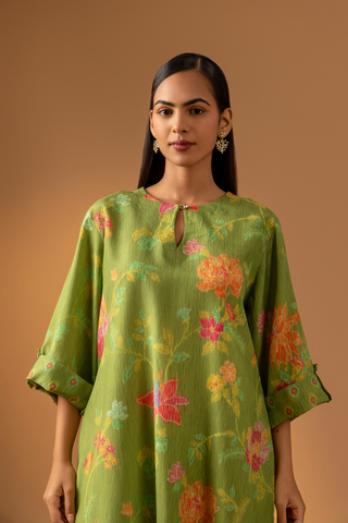 Closeup view of a model stands in a vibrant green co-ord set with floral patterns, paired with matching trousers, against a warm beige background.