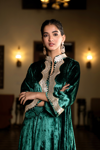 Closeup view of a woman in a green velvet co-ord set traditional outfit stands in a warmly lit room with decorative lighting and patterned flooring.