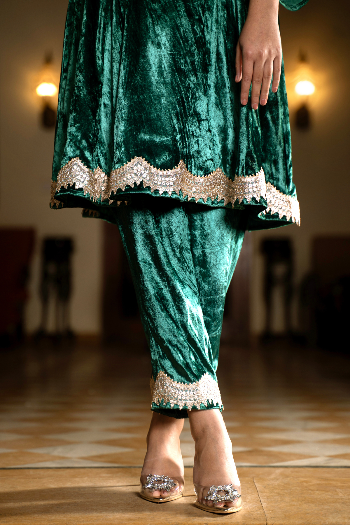A woman stands elegantly in a green velvet traditional outfit with embellished cuffs, showcasing her stylish shoes in a softly lit space.