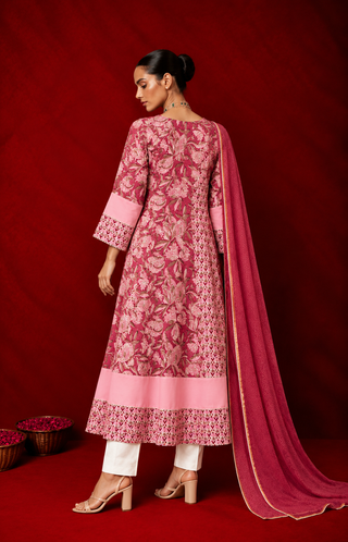 Backside of a woman wearing a pink floral printed anarkali kurta with white pants and a matching dupatta, styled with traditional jewelry, posing against a rich red backdrop.