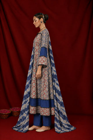 A woman stands in wearing a traditional ajrakh anarkali kurta set consisting of a long printed kurta, blue pants, and a matching dupatta. The kurta has a beige base with intricate red and blue floral patterns, and features solid blue panels on the sleeves and hem.