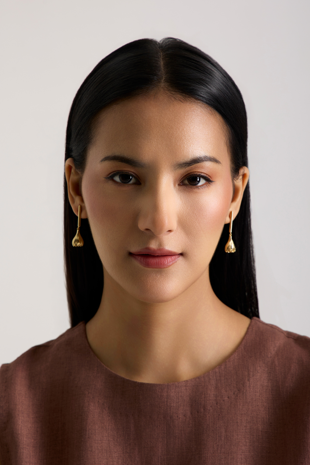 A woman with long black hair wearing small gold dangle earrings and a brown top, photographed against a plain light gray background.