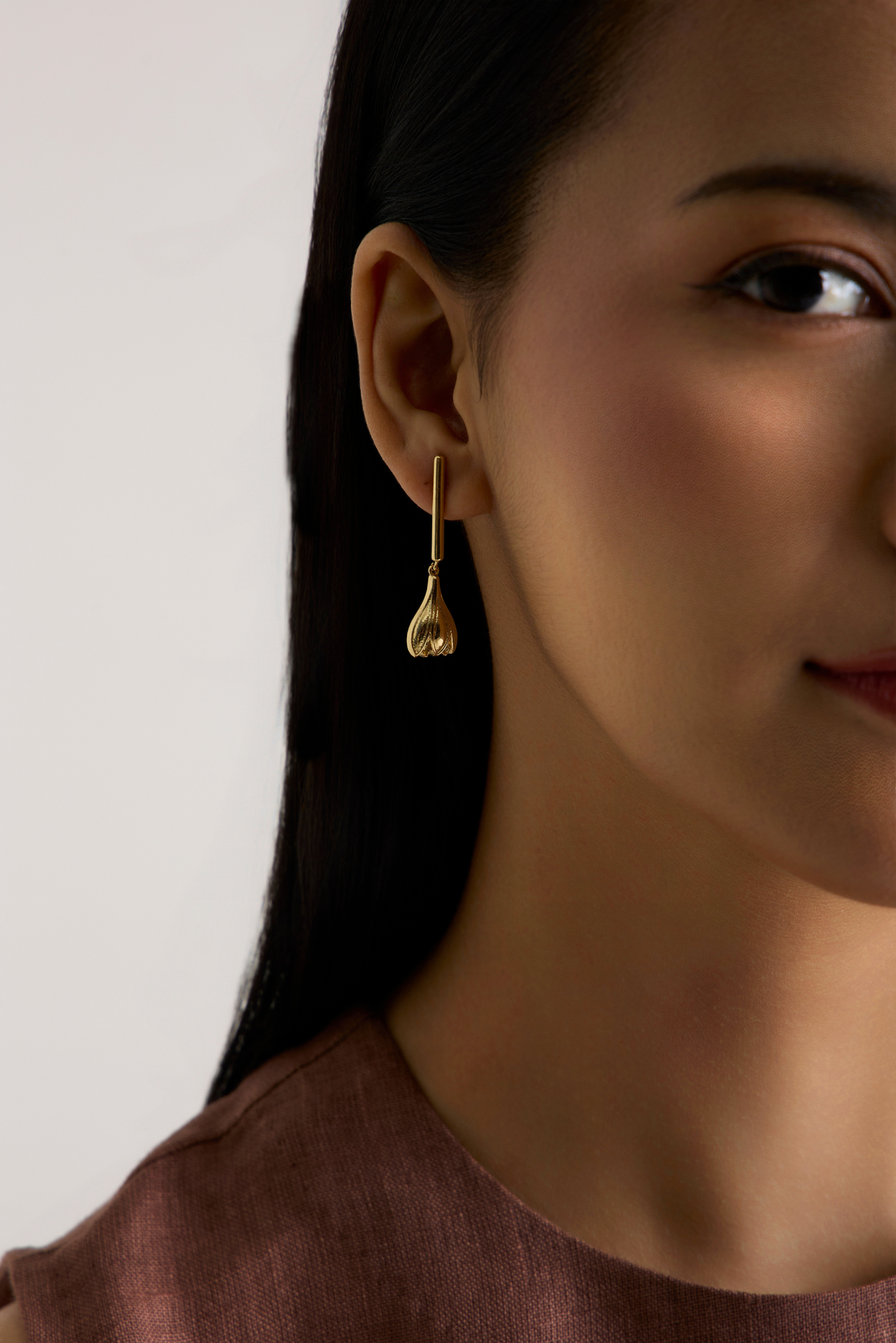 A close-up of a woman wearing a delicate gold dangle earring with a teardrop-shaped drop, shown against a soft light gray background.
