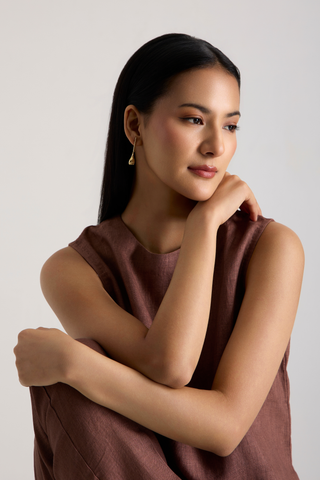 A woman with long black hair wearing a sleeveless brown top and a small gold dangle earring, posing against a light gray background.