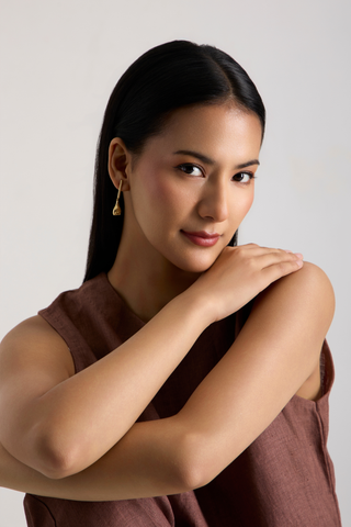 A woman with long dark hair wearing a brown sleeveless top and a small gold dangle earring, posing with her arms crossed against a light gray background.