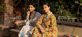 Two women seated outdoors wearing colorful embroidered traditional outfits. The left woman wears a blue, white, and yellow patterned dress, and the right woman wears a bright yellow dress with red and green floral embroidery.