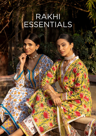 Two women seated outdoors wearing traditional printed ethnic outfits. The right woman wears a bright yellow kurta with red floral motifs and a matching skirt, while the left woman wears a blue, white, and orange patterned outfit.