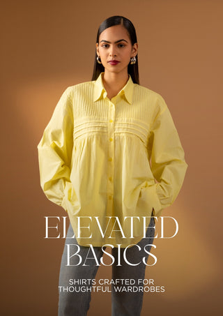 A woman modeling a pale yellow button-up shirt with gathered detailing and long sleeves, paired with gray pants, against a warm brown studio background. Large text at the bottom reads 'ELEVATED BASICS' with smaller text below.