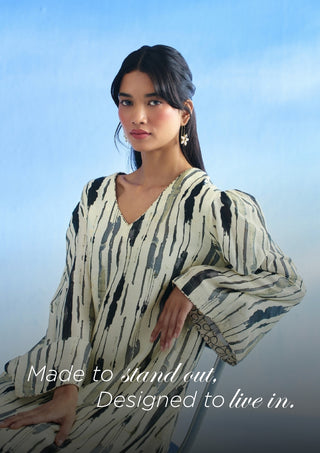 A woman wearing a loose, long-sleeve V-neck c-ord set with an abstract black and gray brushstroke pattern on a cream base, posed against a soft blue gradient background with the text 'Made to stand out, Designed to live in.' overlaid at the bottom.