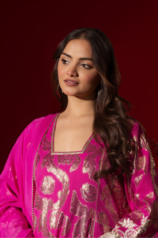 Closeup view of a woman in an elegant pink shimmer kurta set with intricate golden embroidery stands against a vibrant red background.