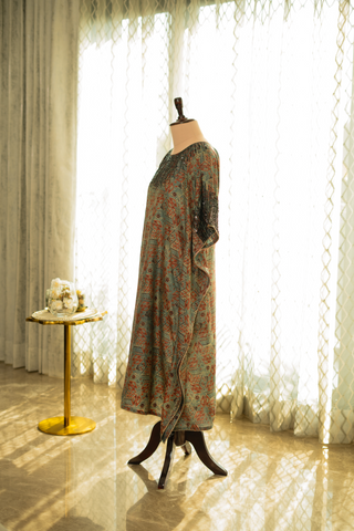 side view of green and rust printed kaftan with intricate pleated detailing and sequin embellishments on the neckline, displayed on a mannequin against a softly lit curtain backdrop.