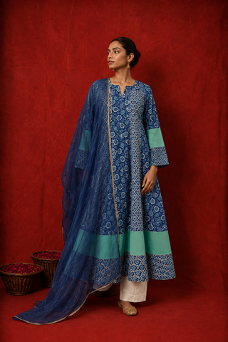 A woman wearing a blue floral printed kurta with white pants and a matching blue dupatta featuring green borders, posed against a rich red backdrop.