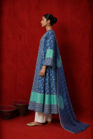 Left side of a woman wearing a blue floral printed kurta with white pants and a matching blue dupatta featuring green borders, posed against a rich red backdrop.