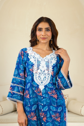 Closeup view of a  woman wearing a blue floral-printed traditional outfit with white embroidered detailing, matching pants, and a patterned blue dupatta, standing in front of an arched alcove in a light-toned interior.